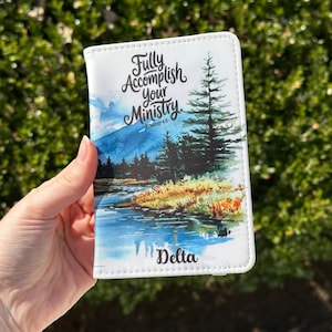May include: White passport holder with a watercolor landscape design featuring a river, trees, and mountains. The text "Fully Accomplish your Ministry" is displayed above the landscape, with "Delta" printed below.