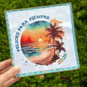 May include: Square art print featuring a beach scene with palm trees, a sunset, and the text "FELICES PARA SIEMPRE" and "2026". The print has a white border with a blue trim and is mounted on a wooden base.