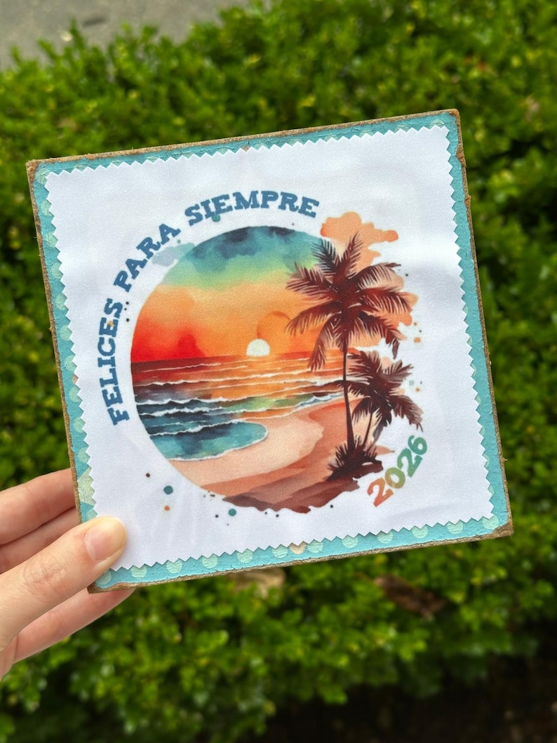 May include: Square art print featuring a beach scene with palm trees, a sunset, and the text "FELICES PARA SIEMPRE" and "2026". The print has a white border with a blue trim and is mounted on a wooden base.