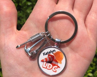 Puede incluir: Un llavero plateado con un dije redondo que presenta un cono de construcción, un casco de obra y el texto "Katelyn LDC". El dije está unido a un llavero plateado con un pequeño destornillador, un martillo y una llave inglesa.