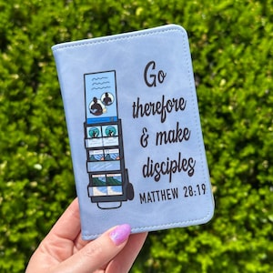 May include: A light blue Bible cover with the text "Go therefore & make disciples" and "Matthew 28:19". The cover features a graphic of a cart with images. The background is a green bush.