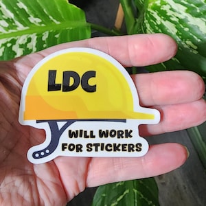 May include: A yellow and orange sticker in the shape of a construction hard hat. The sticker has the letters "LDC" in black and the text "WILL WORK FOR STICKERS" in black. The sticker is held in a hand.