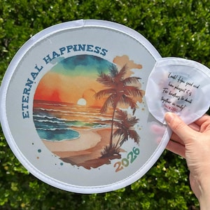 May include: A round, white fan with a watercolor beach scene featuring palm trees, a sunset, and the words "ETERNAL HAPPINESS 2026." A smaller, white, folded item with text is held next to the fan.