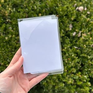 May include: A clear plastic passport holder with a white insert. The holder is rectangular and has a clear border. The background is a green bush.