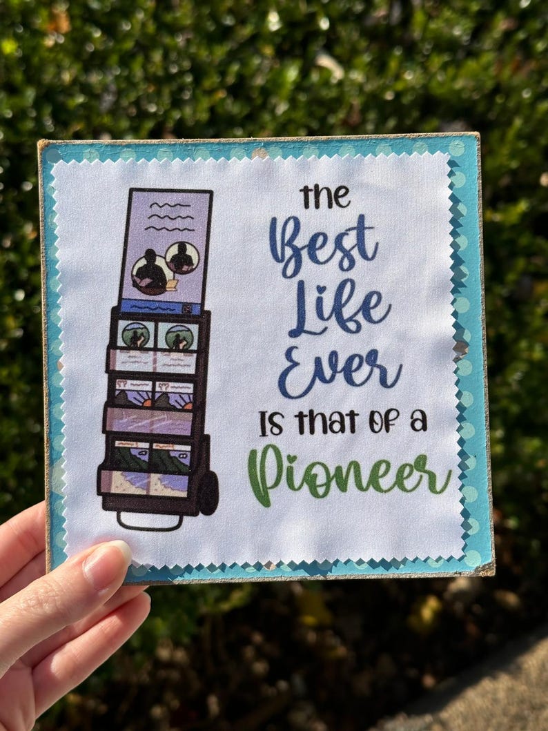 May include: A white sign with a blue border and white polka dots. The sign features a cartoon image of a person with a backpack and a trolley full of luggage. The text on the sign reads "The best life ever is that of a pioneer."