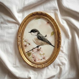 Könnte beinhalten: Ein ovales, goldgerahmtes Kunstwerk mit einer detaillierten Illustration eines kleinen Vogels, der auf einem blühenden Zweig sitzt. Der Vogel hat ein schwarz-weißes Gefieder, und die Blüten sind zartrosa. Der Rahmen hat verzierte Details.