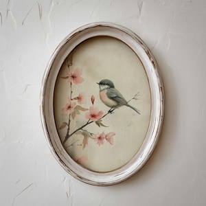 May include: Oval framed artwork featuring a watercolor painting of a bird perched on a flowering branch. The bird has gray and pink plumage. The frame is white with a distressed finish, adding a vintage aesthetic. The artwork is a decorative piece.