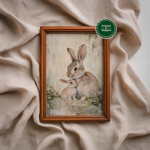 May include: A framed print featuring a mother rabbit and a baby rabbit. The artwork is in soft neutral tones, with the rabbits in shades of brown and beige. The frame is a warm brown color. The print has a sticker that says "Printed & Shipped".