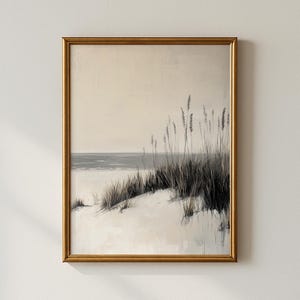 May include: A framed art print depicting a serene beach scene. The artwork features a sandy dune with tall, wispy grasses in shades of gray and black, set against a muted sky and ocean. The frame is a warm gold color.