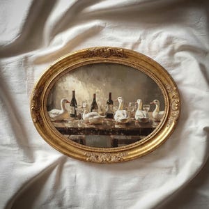 May include: An oval gold-framed painting shows white geese at a table with wine bottles and glasses. The artwork has a vintage style, with soft lighting and a detailed, textured appearance. The frame has an ornate design.
