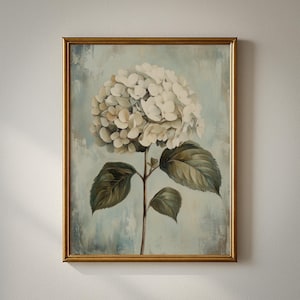 May include: A framed floral print featuring a large white hydrangea bloom. The artwork has a gold-colored frame and a soft blue and white background. The leaves are green and the stem is brown.