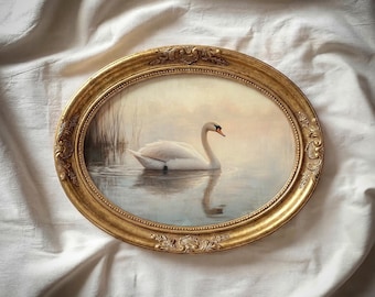 May include: An oval framed painting depicting a swan on a lake. The swan is white with a red beak, and its reflection is visible in the water. The gold frame has decorative detailing. The background shows a soft landscape.