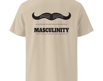 Puede incluir: Una camiseta beige de manga corta con un gráfico negro de un bigote estilizado sobre la palabra MASCULINITY en una fuente serif negrita. El texto está encerrado entre dos líneas horizontales negras, creando un diseño centrado.