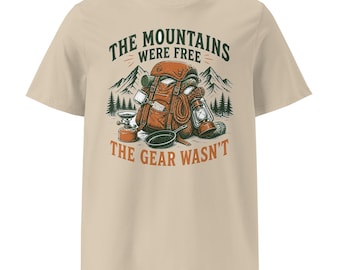 May include: A beige short-sleeve t-shirt featuring a graphic of a brown backpack, a camping stove, a frying pan, and a lantern in front of mountains. The text reads THE MOUNTAINS WERE FREE THE GEAR WASN'T in dark green and orange lettering.
