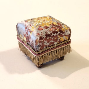 May include: A small, square footstool with a patterned fabric top in silver, gold, and burgundy. The stool has a burgundy trim with gold fringe and sits on four small wooden legs. The overall design is ornate and decorative.