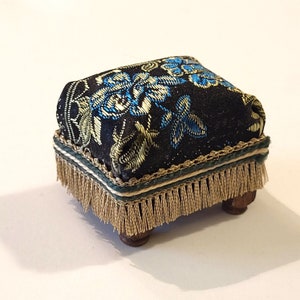 May include: A small, square pincushion with a black fabric top featuring a floral design in blue and gold. The pincushion has a green and gold trim, gold fringe, and sits on four small wooden feet.