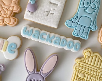 May include: A set of decorated sugar cookies featuring various characters and designs. The cookies are decorated with blue, yellow, pink, and white icing. The cookies include a dog, a bunny, a house, a bone, and a banner that says "Wackadoo".