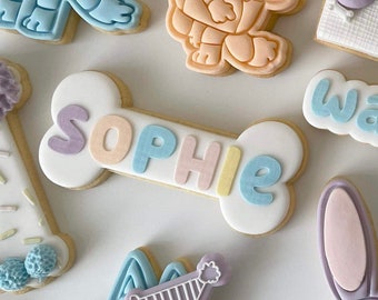 May include: A collection of colorful, decorated sugar cookies. The cookies are shaped like a bone, a dog, a hat, and a cloud. The bone cookie has the name "Sophie" written on it in pastel colors.