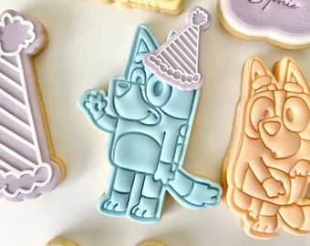 May include: A set of decorated sugar cookies featuring a blue cartoon dog wearing a purple and white party hat, a purple and white party hat, a yellow cookie with the word "Happy" on it, a purple cookie with the word "Sophie" on it, and a light orange cartoon dog.