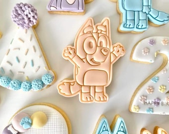 May include: A set of decorated sugar cookies featuring a cartoon dog, a party hat, a number two, and a paw print design. The cookies are decorated with pastel colors and sprinkles.