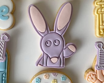 May include: A set of decorated sugar cookies featuring a purple bunny with pink ears, a blue cookie with a party hat, a yellow cookie with a fire hydrant, and a white cookie with paw prints.
