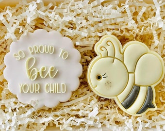 Puede incluir: Dos galletas de azúcar decoradas, una con el texto "So proud to bee your child" y la otra con un diseño de abeja blanca. Las galletas están sobre una cama de papel blanco triturado.