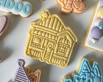 May include: A set of decorated sugar cookies featuring a yellow house, a blue dog, a purple party hat, and a white cookie with the word "ADOO" in blue lettering.