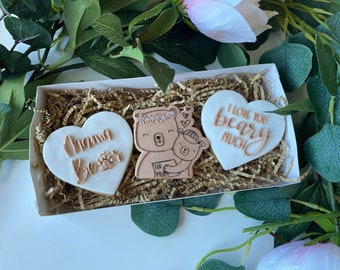 Puede incluir: Una caja de regalo llena de tres galletas con forma de corazón decoradas con glaseado marrón. Una galleta dice "Mama Bear" con una huella de pata, otra dice "I love you bearly much", y la tercera presenta un oso de dibujos animados y una cría.