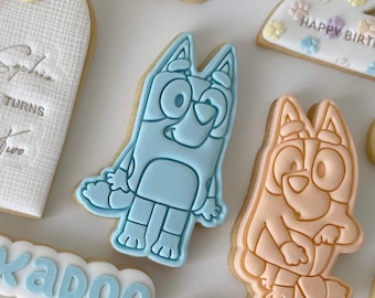 May include: Two decorated sugar cookies featuring a blue and an orange cartoon dog. The cookies are decorated with icing and have a simple outline.
