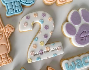 May include: A collection of decorated sugar cookies. The centerpiece is a number 2 cookie, white with pastel paw prints and "HAPPY BIRTHDAY" text. Other cookies feature cartoon dog characters and paw prints.