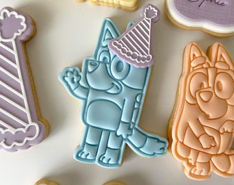 May include: A set of decorated sugar cookies featuring a blue dog wearing a purple and white party hat, an orange dog, and a purple and white party hat with stripes.