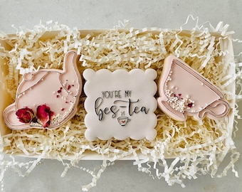 Puede incluir: Una caja de regalo llena de tres galletas de azúcar decoradas en rosa y blanco. Las galletas tienen forma de tetera, taza de té y un cuadrado con el texto "You're my bes-tea" en letras negras.