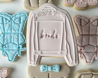 Puede incluir: Un conjunto de galletas de azúcar decoradas con varios diseños, que incluyen un patrón floral azul y blanco, una chaqueta vaquera rosa con la palabra "bride" escrita en ella y una galleta marrón con una pajarita.