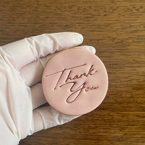 May include: A pink round cookie with the words "Thank You" written in cursive on it.