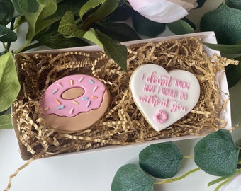 Puede incluir: Una caja de regalo blanca llena de papel de seda marrón contiene dos galletas de azúcar decoradas. Una galleta tiene forma de donut con glaseado rosa y chispas de colores. La otra galleta tiene forma de corazón y dice "No sé qué haría sin ti".
