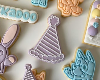 May include: A set of decorated sugar cookies featuring a purple party hat, a blue dog, a pink bunny, and a cookie with the word "skadoodle" in blue icing.