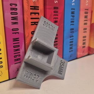 Puede incluir: Soporte gris para lentes de cámara, impreso en 3D, con medidas de 25 mm, 35 mm y 50 mm. El soporte está en ángulo y tiene una abertura cuadrada. Lomos de libros coloridos en el fondo.