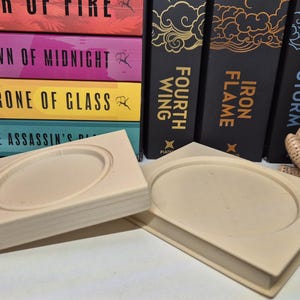 May include: Two beige rectangular soap dishes with an oval indentation. Behind them are books with titles including "Heir of Fire" and "Crown of Midnight." A small figurine of a person reading a book is also present.