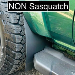 May include: Close-up of a green vehicle's side, featuring a black mud flap and a portion of a large tire. The tire has a rugged tread pattern. The text "NON Sasquatch" is displayed in a white font on a black background.