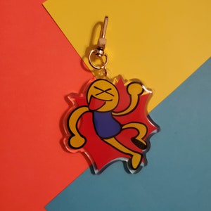 May include: A clear acrylic keychain featuring a cartoon figure with a yellow head, black eyes, and a red tongue. The figure is wearing a blue shirt and is set against a red burst design. The keychain has a gold-colored clasp.
