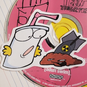 May include: Stickers featuring characters from the animated series, including a cartoon milkshake with a pink straw and a bomb with a radiation symbol. The stickers are on a pink CD with text from the show.