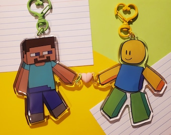 May include: Two character keychains, one of a green and blue Minecraft character holding hands with a yellow and blue Roblox character. Both characters are holding a pink heart charm.