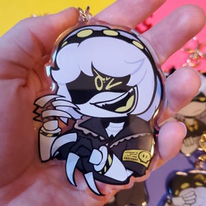 May include: A clear acrylic keychain featuring a cartoon character with white hair, black eyes, and a yellow skull on a black jacket. The character is holding a weapon with sharp teeth.