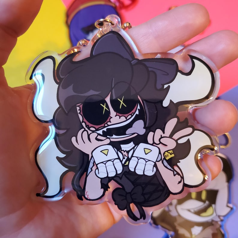 May include: A clear acrylic keychain featuring a cartoon character with black hair, a black dress, and a white bow. The character has a wide smile, red eyes, and white X's over the eyes. The character is holding up two fingers in a peace sign.