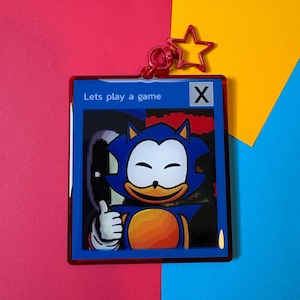 May include: A square acrylic keyring featuring a blue character with a white face and orange belly, giving a thumbs up. The design includes the text 'Lets play a game' and a black 'X' in a white box. It has a red star-shaped metal clasp at the top.