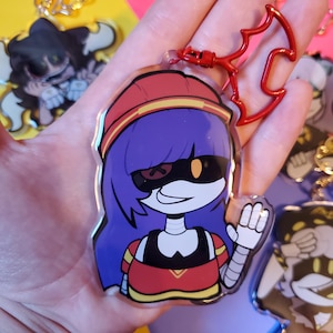 May include: A red, white, and yellow cartoon character with purple hair and a black and yellow mask. The character is wearing a red and yellow shirt and has a robotic arm. The character is holding up a hand in a wave. The character is a keychain.