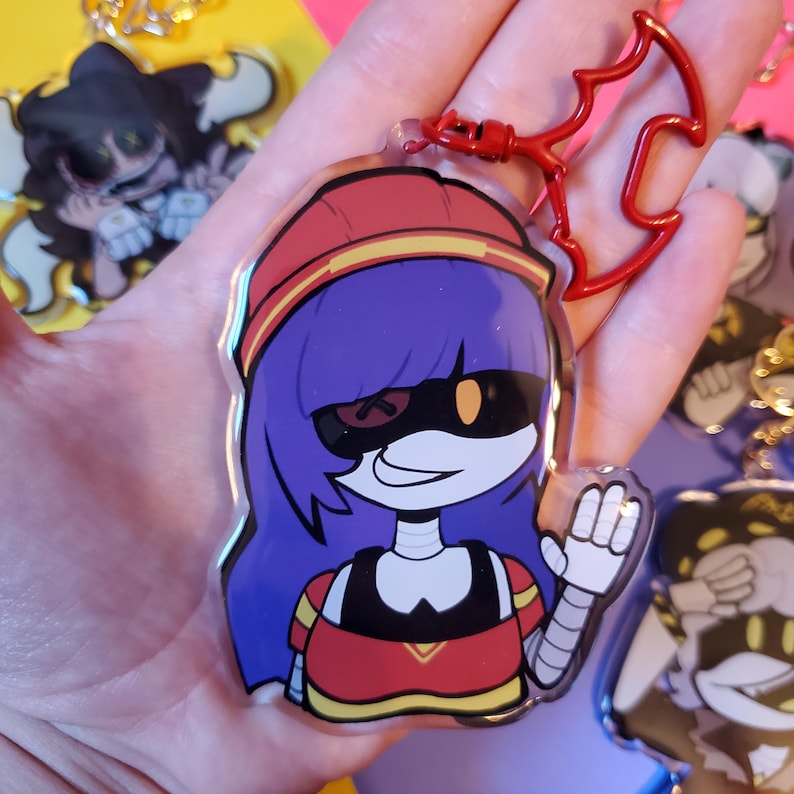 May include: A red, white, and yellow cartoon character with purple hair and a black and yellow mask. The character is wearing a red and yellow shirt and has a robotic arm. The character is holding up a hand in a wave. The character is a keychain.