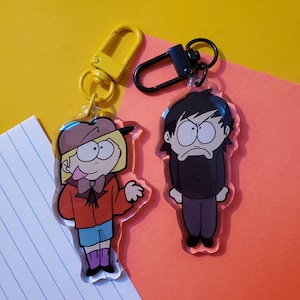 May include: Two cartoon character keychains. One keychain features a girl with blonde hair wearing a red jacket, blue pants, and a brown hat. The other keychain features a boy with black hair wearing a black shirt and pants. Both keychains are made of clear acrylic.