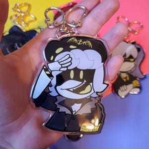 May include: A black and white cartoon character keychain with a yellow and black hat. The character is covering one eye with their hand and has a wide smile. The keychain is attached to a silver key ring.