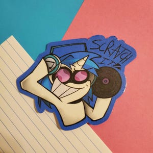 May include: A sticker featuring a cartoon unicorn DJ with blue hair, a horn, and pink sunglasses. The unicorn is holding a record and wearing headphones. The text "SCRATCH IT" is above the unicorn.